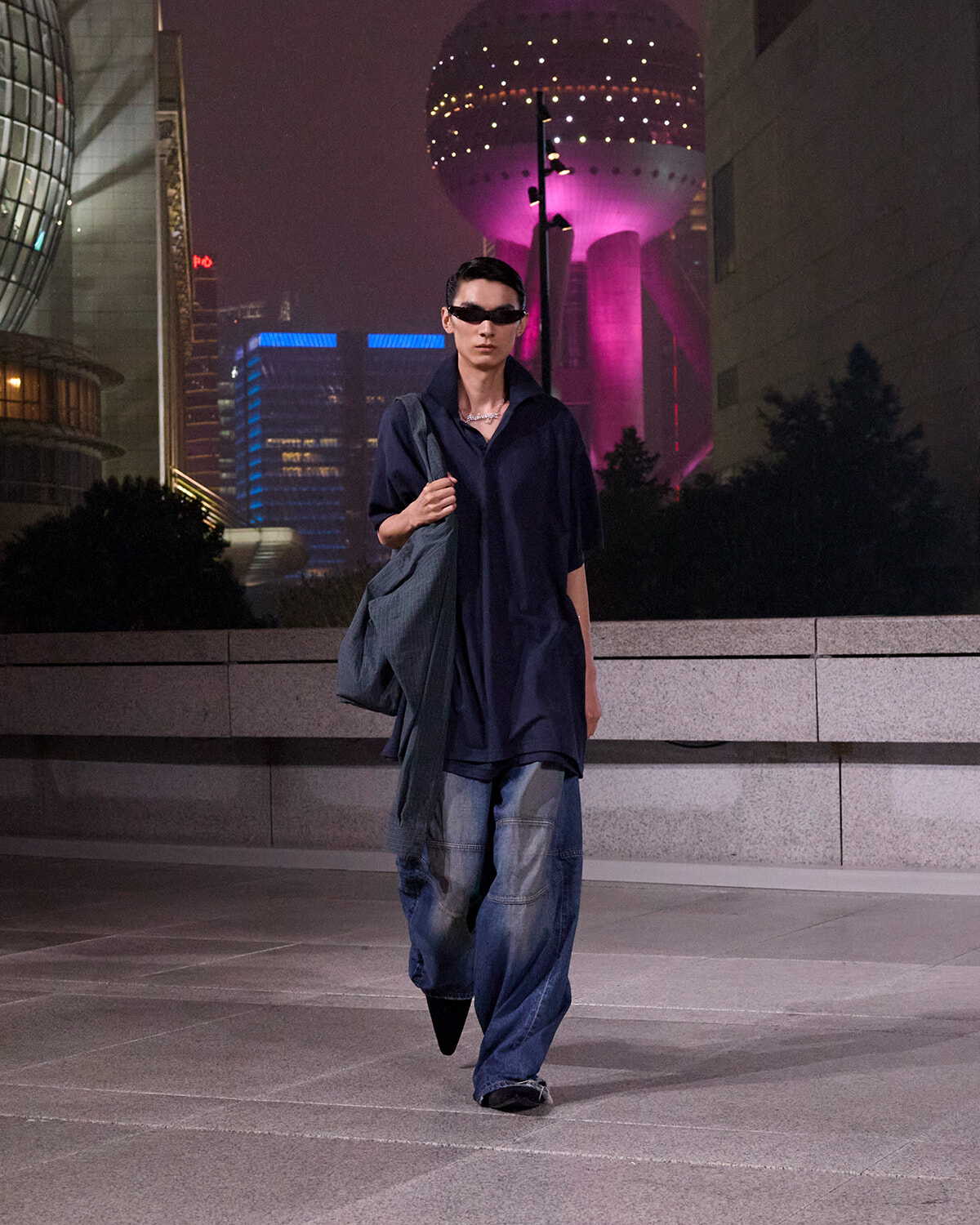 Man in a dark oversized shirt and baggy jeans walks at night in a neon cityscape, carrying a gray shoulder bag.