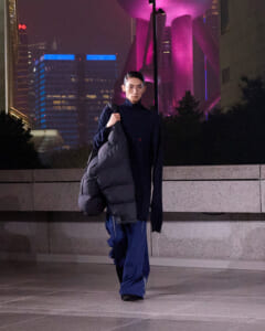 Person in dark clothing walks along a lit city sidewalk at night, carrying a large gray puffer jacket over their shoulder with purple-lit buildings in the background.