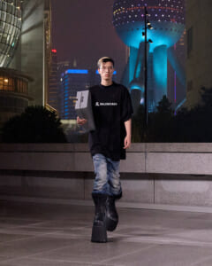 Young man in a black Balenciaga T-shirt walks on a nighttime city sidewalk, carrying two gray boxes, wearing faded jeans and chunky black boots, neon buildings in the background.
