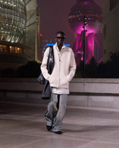 Man wearing an oversized cream jacket and baggy jeans, with a black shoulder bag, walking at night in a neon-lit city skyline backdrop.