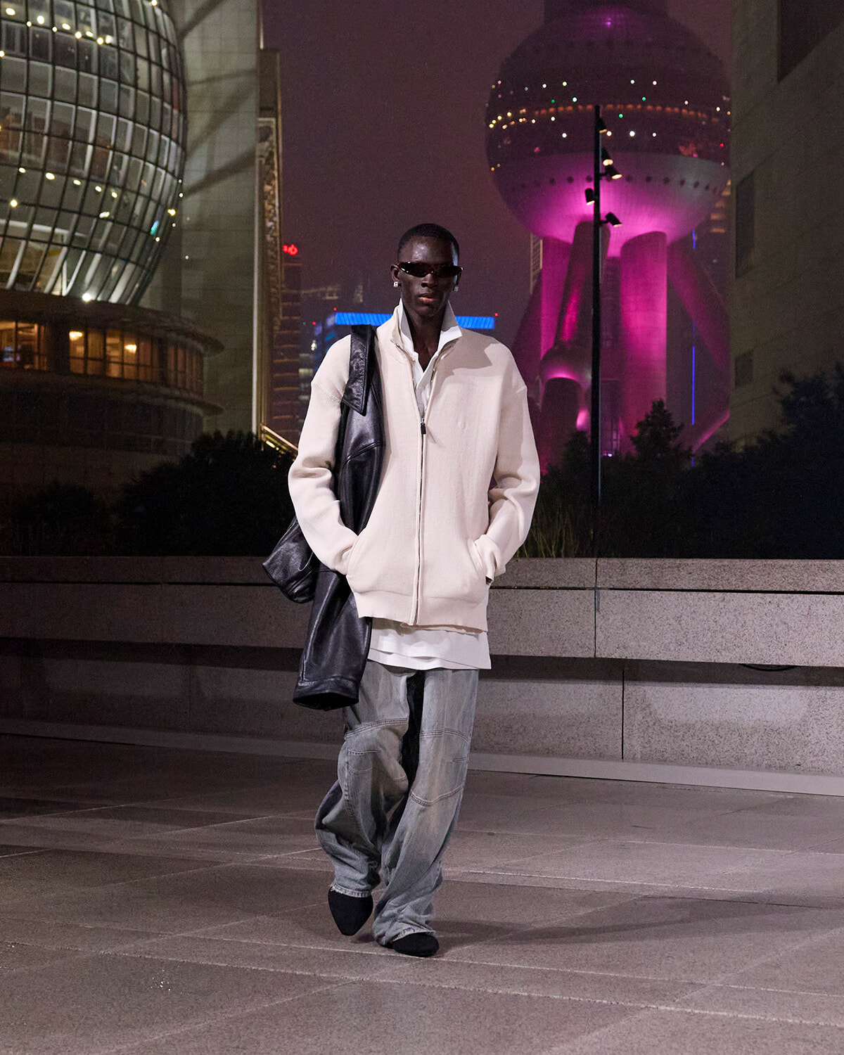 Man wearing an oversized cream jacket and baggy jeans, with a black shoulder bag, walking at night in a neon-lit city skyline backdrop.