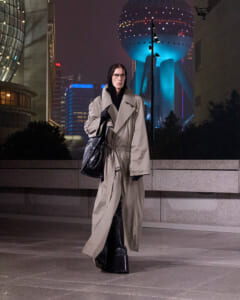 Person in a long beige trench coat and black boots walks on a city sidewalk at night, with futuristic neon buildings behind him/her.