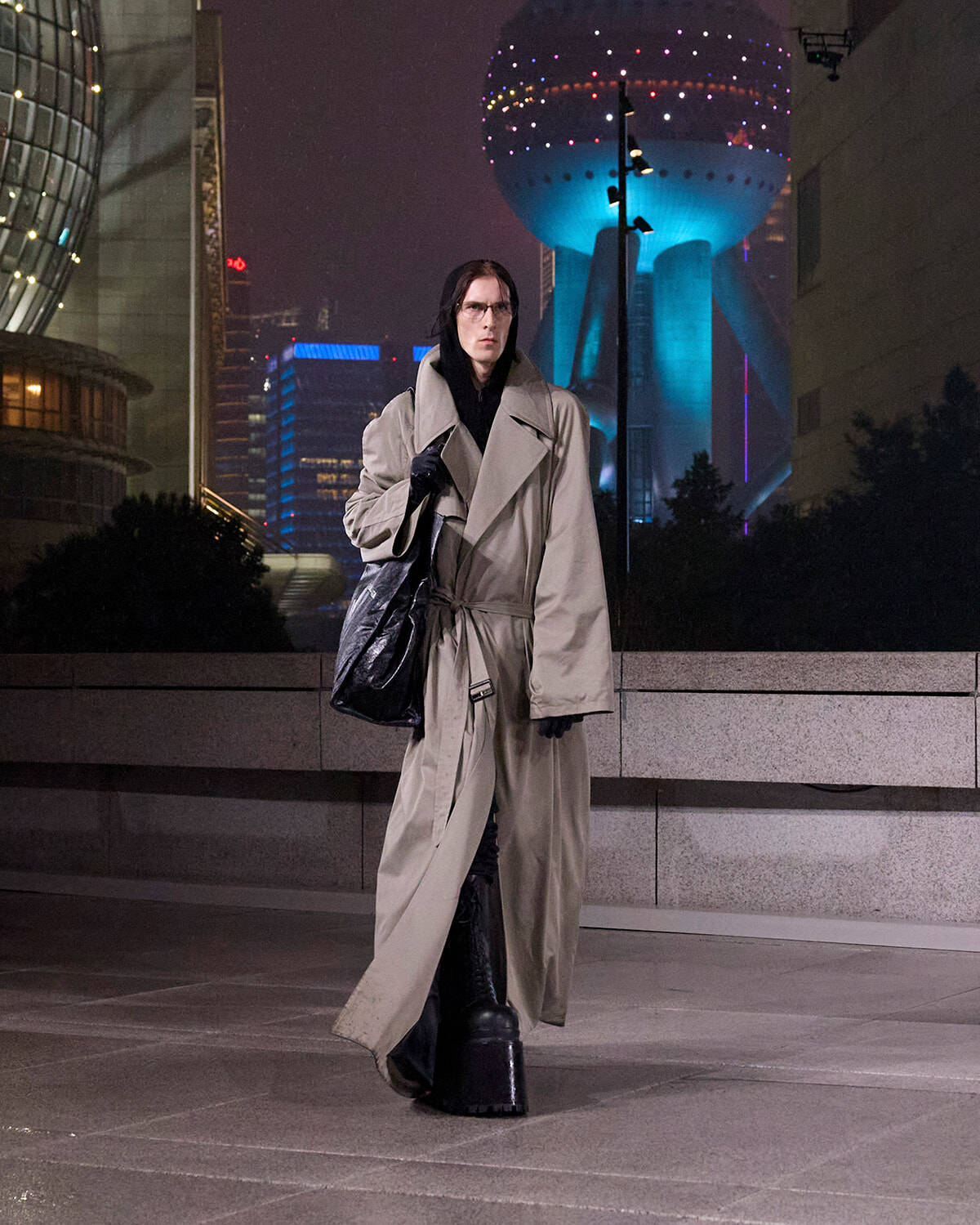 Person in a long beige trench coat and black boots walks on a city sidewalk at night, with futuristic neon buildings behind him/her.