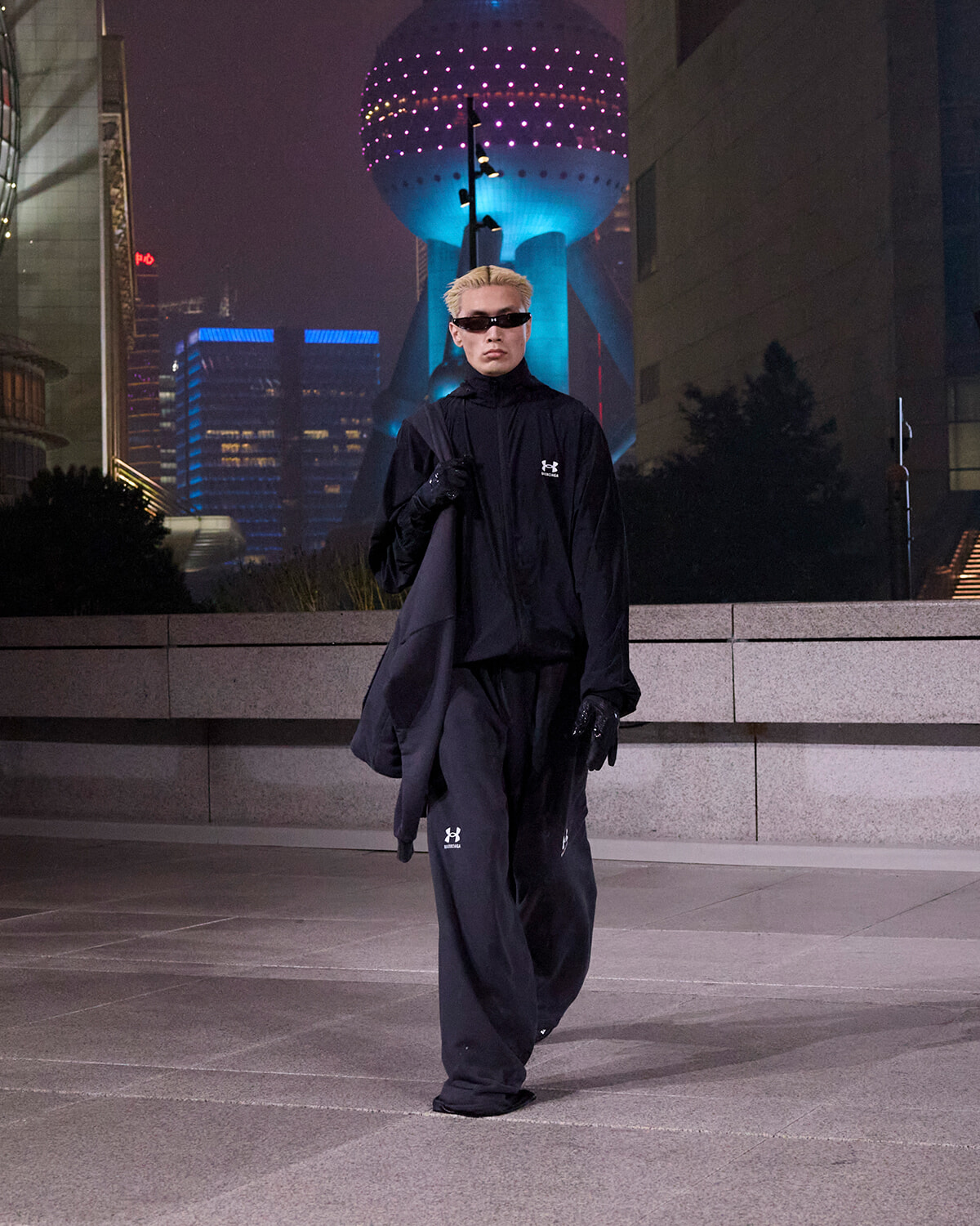 Person in black oversized outfit and sunglasses walks across a nighttime city square with a neon-lit spherical tower in the background.