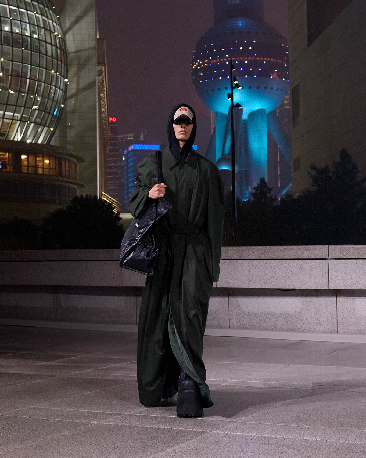 Person walking in a dark green trench coat and hood, carrying a black bag, in a nighttime urban plaza with neon-lit buildings behind.
