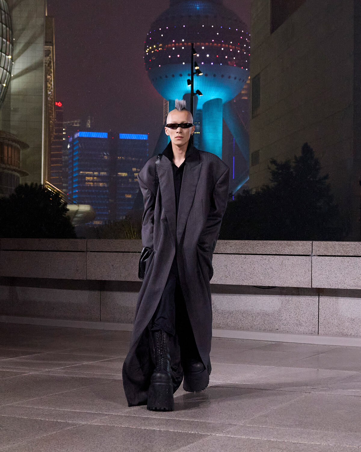 Person with gray mohawk in an oversized black coat and sunglasses walks on a city street at night, with a neon-lit sphere building in the background.