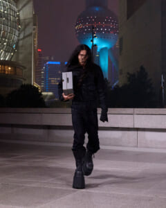 Person with long dark hair in black outfit and chunky boots walking at night, carrying a white box against a city plaza backdrop.