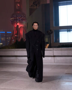 Person wearing a long black double-breasted coat walking on a tiled plaza at night, with a lit cityscape in the background.