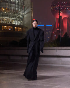 Person in an oversized black suit stands on a night city plaza with futuristic glass buildings behind him/her, creating a moody urban scene.