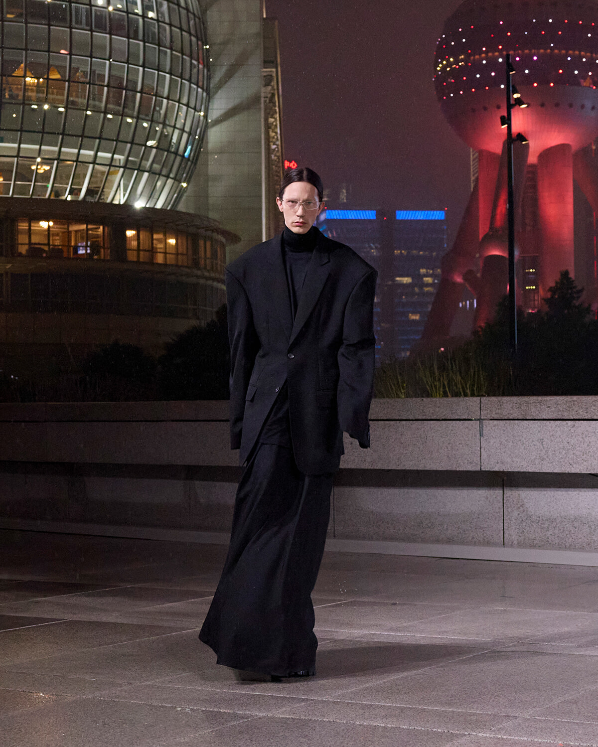 Person in an oversized black suit stands on a night city plaza with futuristic glass buildings behind him/her, creating a moody urban scene.