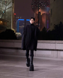 Person wearing an oversized black coat and tall boots walks toward the camera on a nighttime city plaza with illuminated buildings in the background.