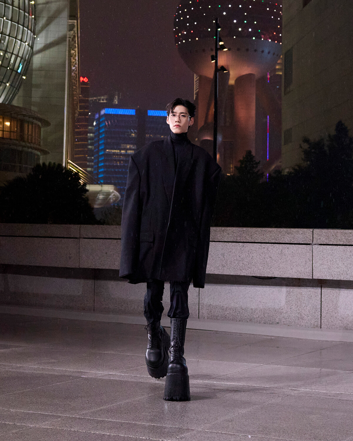 Person wearing an oversized black coat and tall boots walks toward the camera on a nighttime city plaza with illuminated buildings in the background.