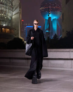 Model in an oversized black coat and boots walking on a nighttime city street with futuristic buildings behind.