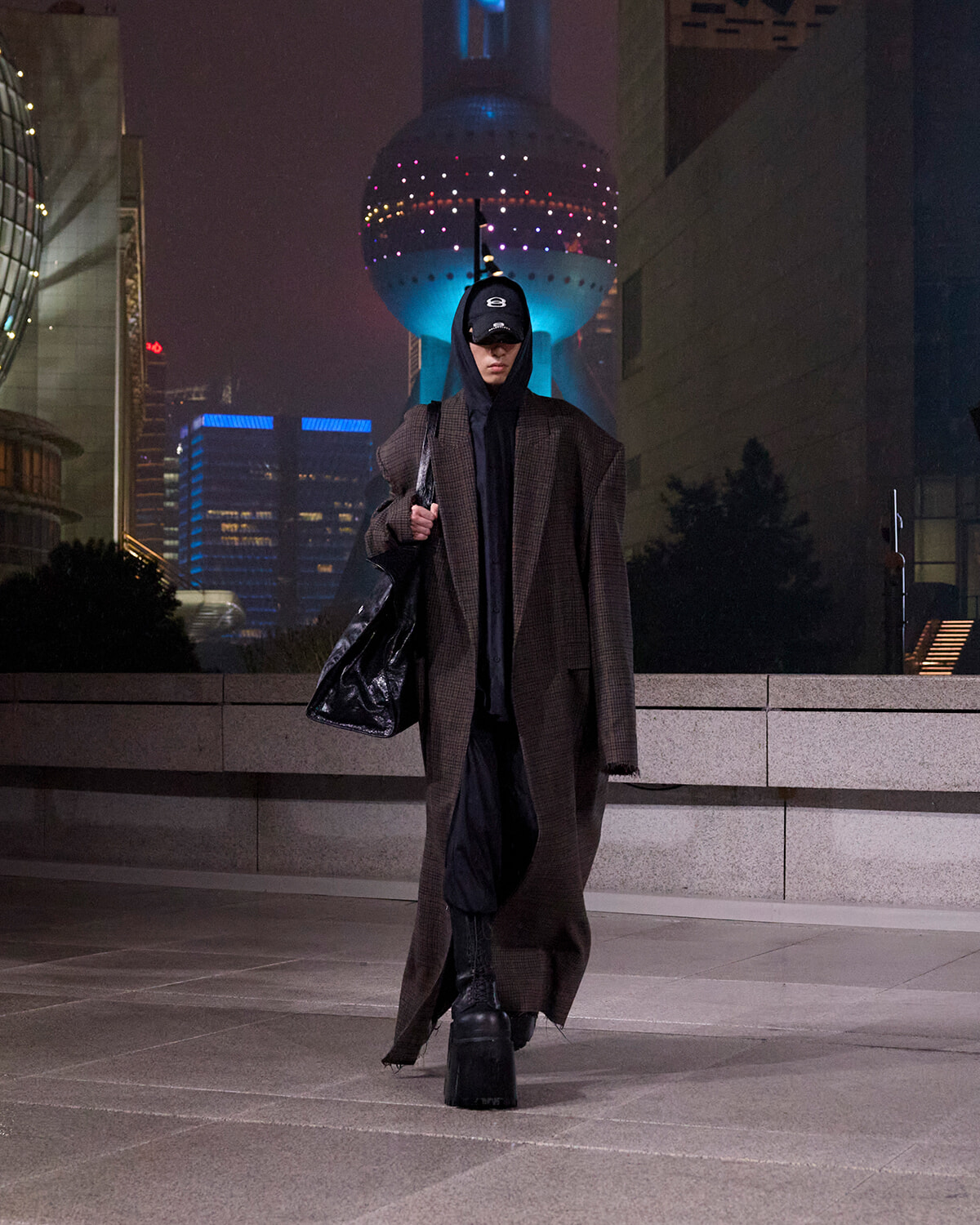 Person wearing a long brown coat, hoodie, and cap carries a large black bag while walking at night in a modern city plaza with a lit, spherical building in the background.