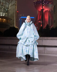 Person wearing an oversized pale blue floral gown with a large front bow, walking on a city sidewalk at night.