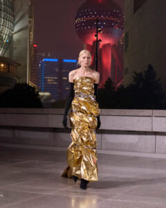 Model wearing a gold metallic strapless gown with long black gloves walking on a city pavement at night, neon lights in the background.