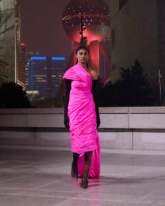Woman in a bright pink, one-shoulder draped gown and black opera gloves posing outdoors at night with city skyline and illuminated tower in the background.