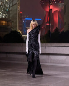 Fashion model in a one-shoulder, black draped gown with long white gloves, standing on a city plaza at night with neon lights behind.