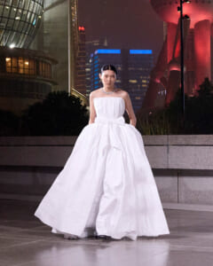 Woman in a strapless white ball gown posing on a city plaza at night with illuminated skyscrapers behind her.