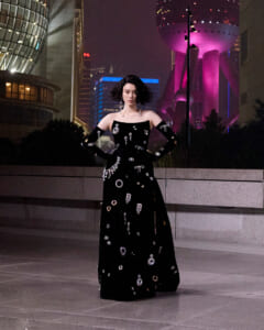 Fashion model in an off-shoulder black floor-length dress with silver embellishments posing with hands on hips at night in a city plaza.