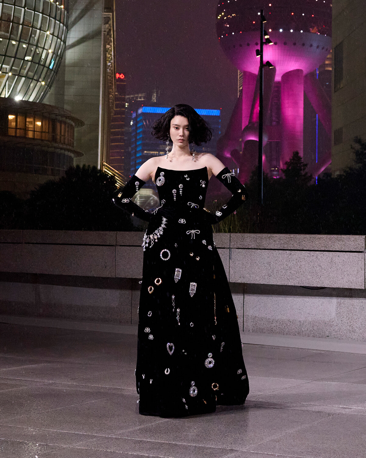 Fashion model in an off-shoulder black floor-length dress with silver embellishments posing with hands on hips at night in a city plaza.