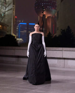 Person wearing a long black strapless gown and white elbow-length gloves stands in an outdoor urban plaza at night, with modern city buildings in the background.
