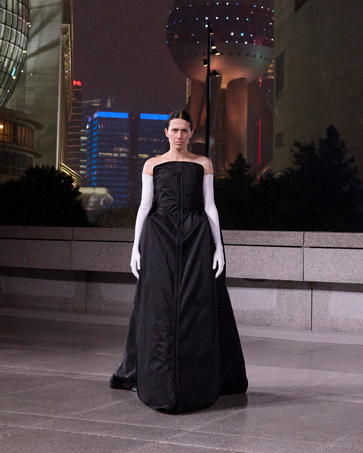 Person wearing a long black strapless gown and white elbow-length gloves stands in an outdoor urban plaza at night, with modern city buildings in the background.