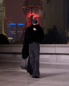 Person in a black fur coat and glittery wide-leg pants walks at night in a city plaza, carrying a metallic mesh bag with a large illuminated sphere sculpture behind them.