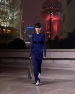 Woman in a royal blue, long-sleeve dress walks along a concrete plaza at night, carrying a metallic chain bag; futuristic city lights in the background.