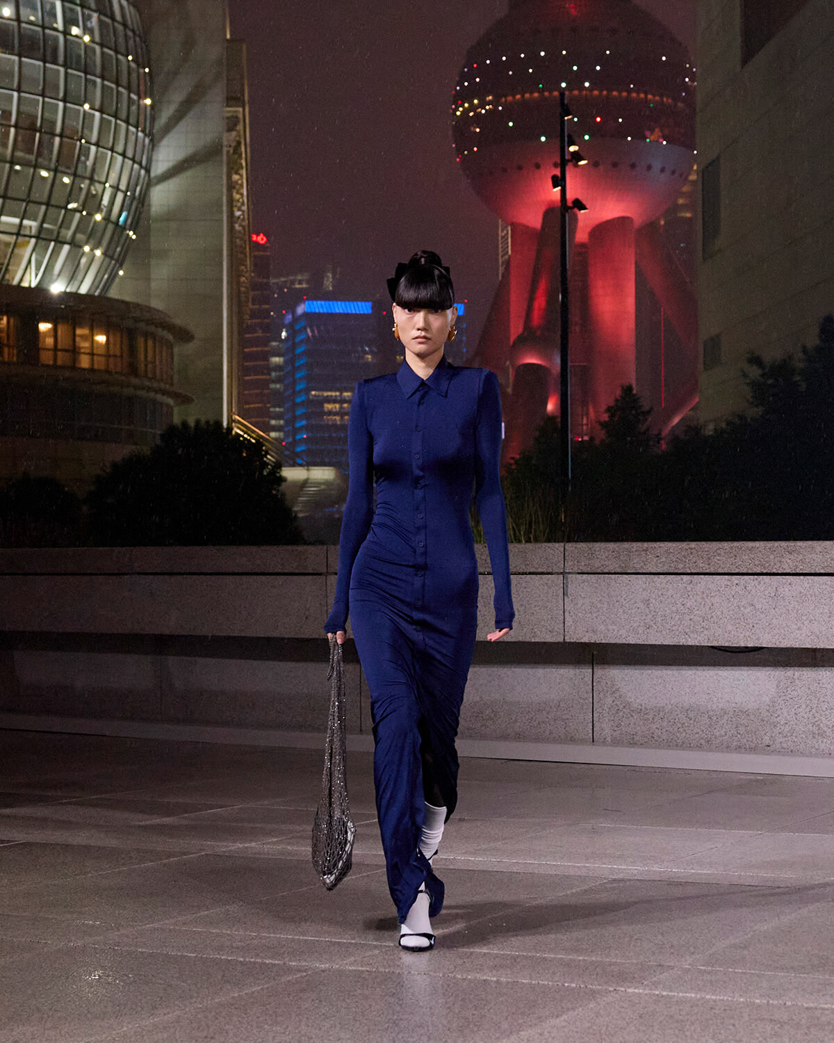 Woman in a royal blue, long-sleeve dress walks along a concrete plaza at night, carrying a metallic chain bag; futuristic city lights in the background.