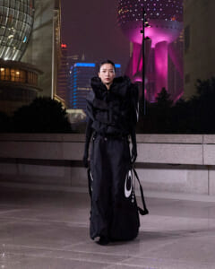 A person wearing an oversized black avant-garde outfit stands outdoors at night in a city square, with a large black bag featuring a white circular logo beside them and purple-lit skyscrapers in the background.