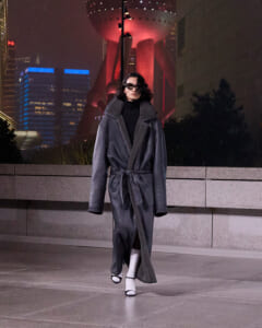 Person in a long dark shearling coat and sunglasses stands on a city plaza at night, with neon-lit buildings in the background.