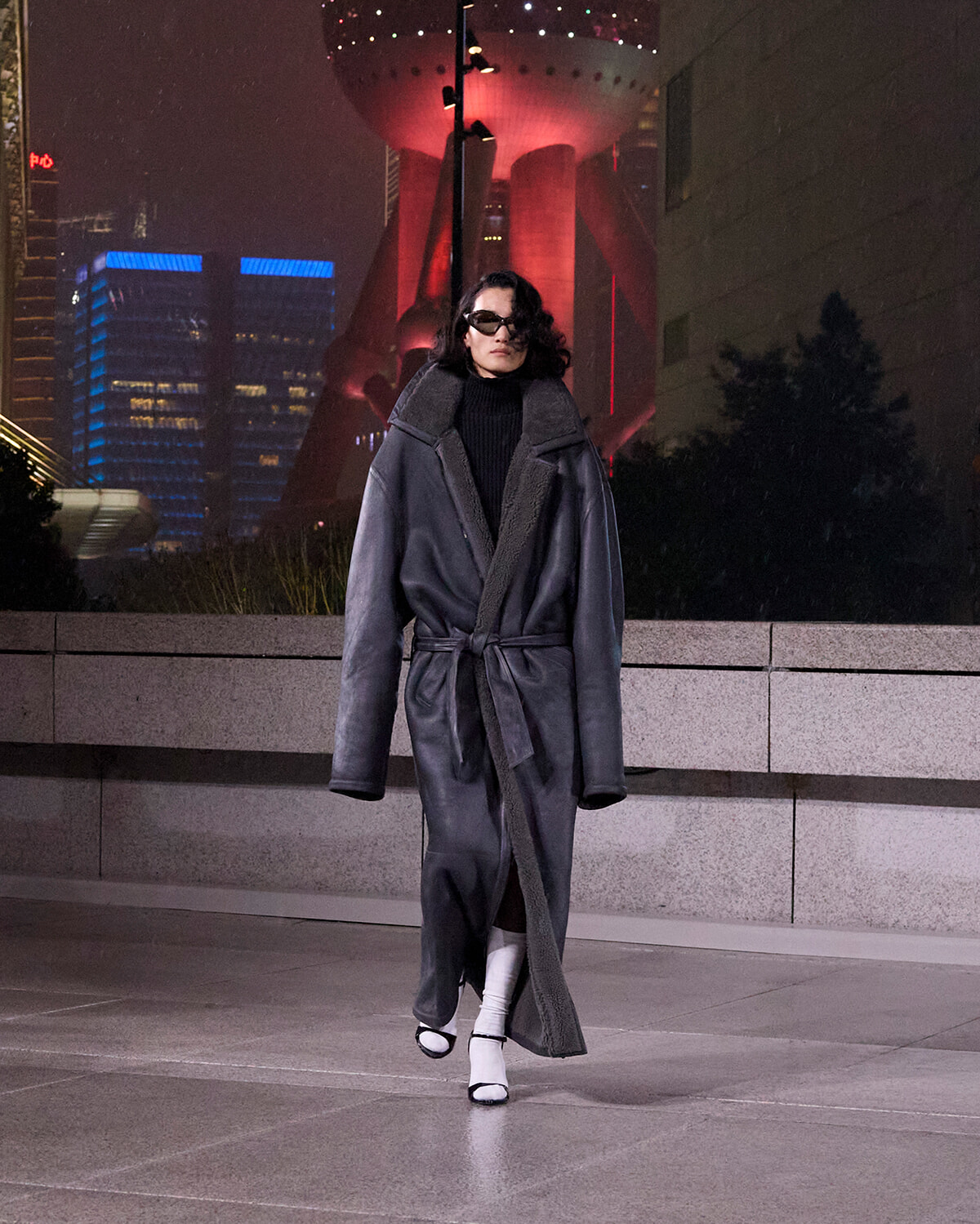 Person in a long dark shearling coat and sunglasses stands on a city plaza at night, with neon-lit buildings in the background.