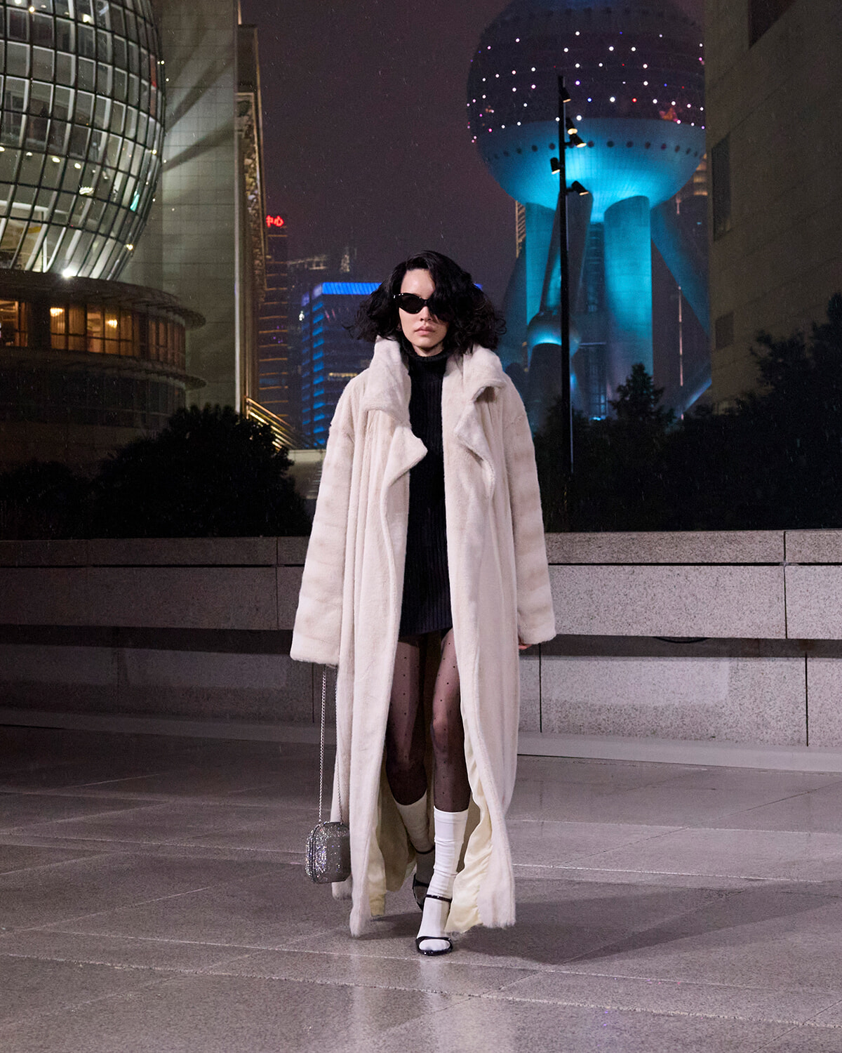 Woman in a long cream fur coat stands in a night-time city plaza with blue-lit towers behind her.