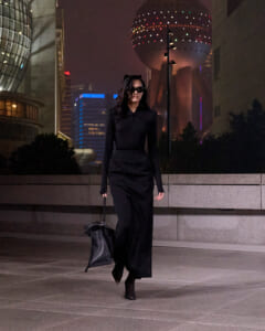 Woman in an all-black outfit and sunglasses walking on a city sidewalk at night, carrying a black bag.