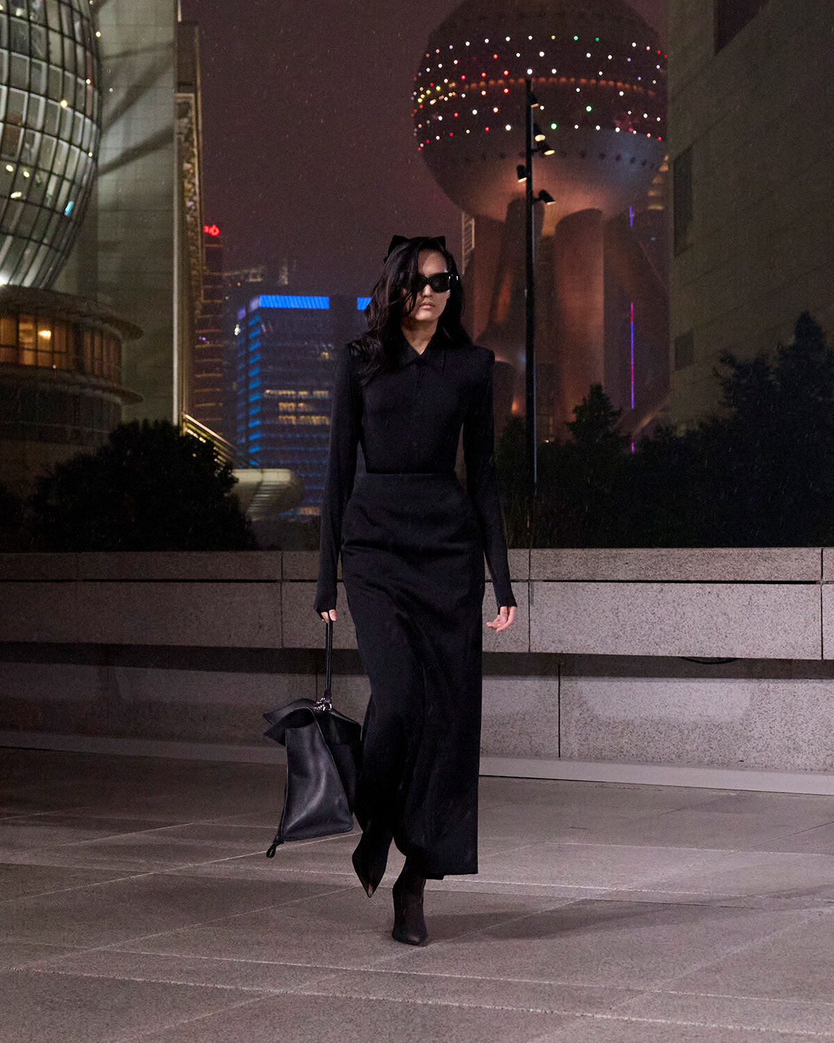 Woman in an all-black outfit and sunglasses walking on a city sidewalk at night, carrying a black bag.