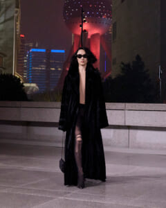 Woman in a long black fur coat and sunglasses walking on a city sidewalk at night, neon skyline in the background, carrying a small clutch.