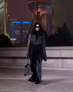 Person wearing an oversized black leather coat and sunglasses walks at night in a cityscape, carrying a gray handbag.
