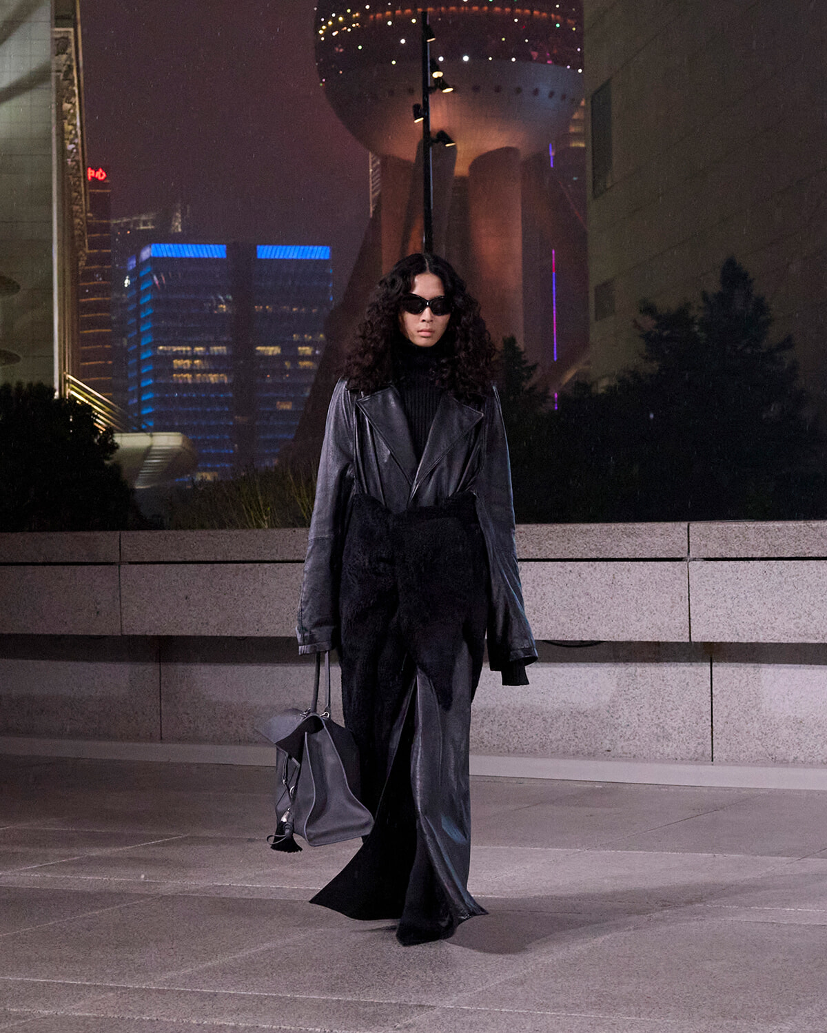Person wearing an oversized black leather coat and sunglasses walks at night in a cityscape, carrying a gray handbag.