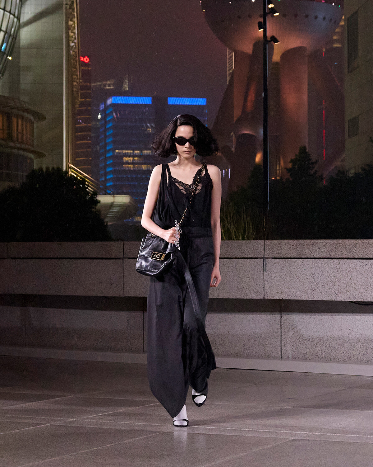 Woman in a black lace top and wide-leg pants walks at night in a city plaza, carrying a black quilted shoulder bag and wearing sunglasses.