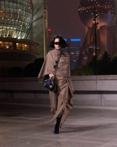 Woman in a brown plaid cape and matching pants, black ankle boots, and a black quilted bag walks at night in a city plaza.