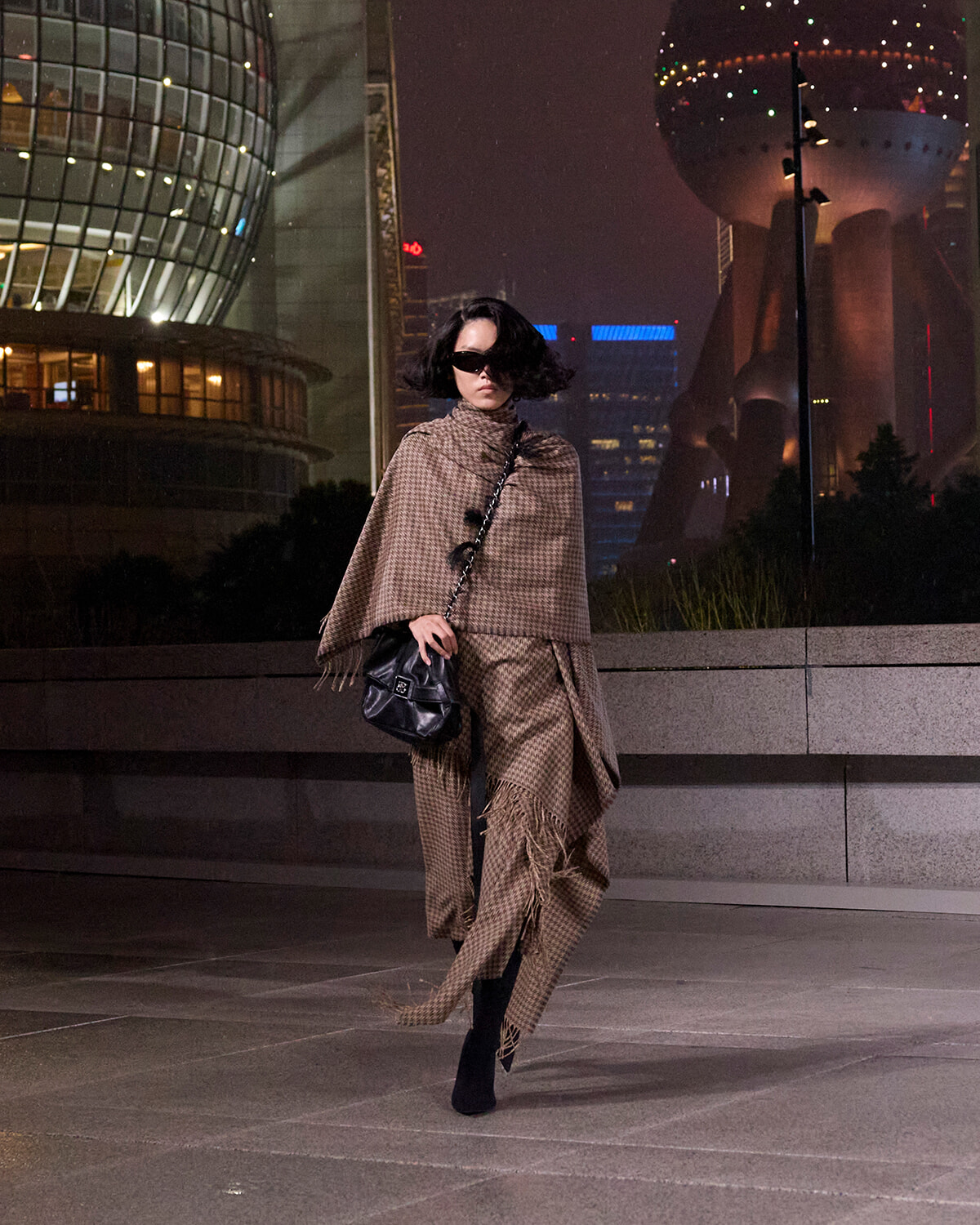 Woman in a brown plaid cape and matching pants, black ankle boots, and a black quilted bag walks at night in a city plaza.