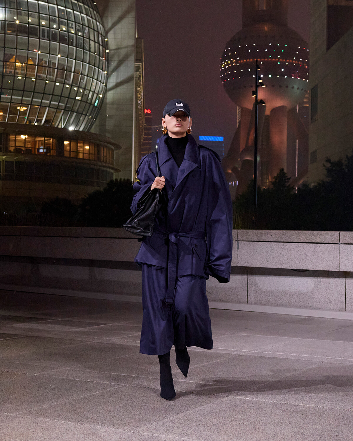 Person in oversized navy trench coat and cap walks at night in a city plaza with futuristic buildings behind them.