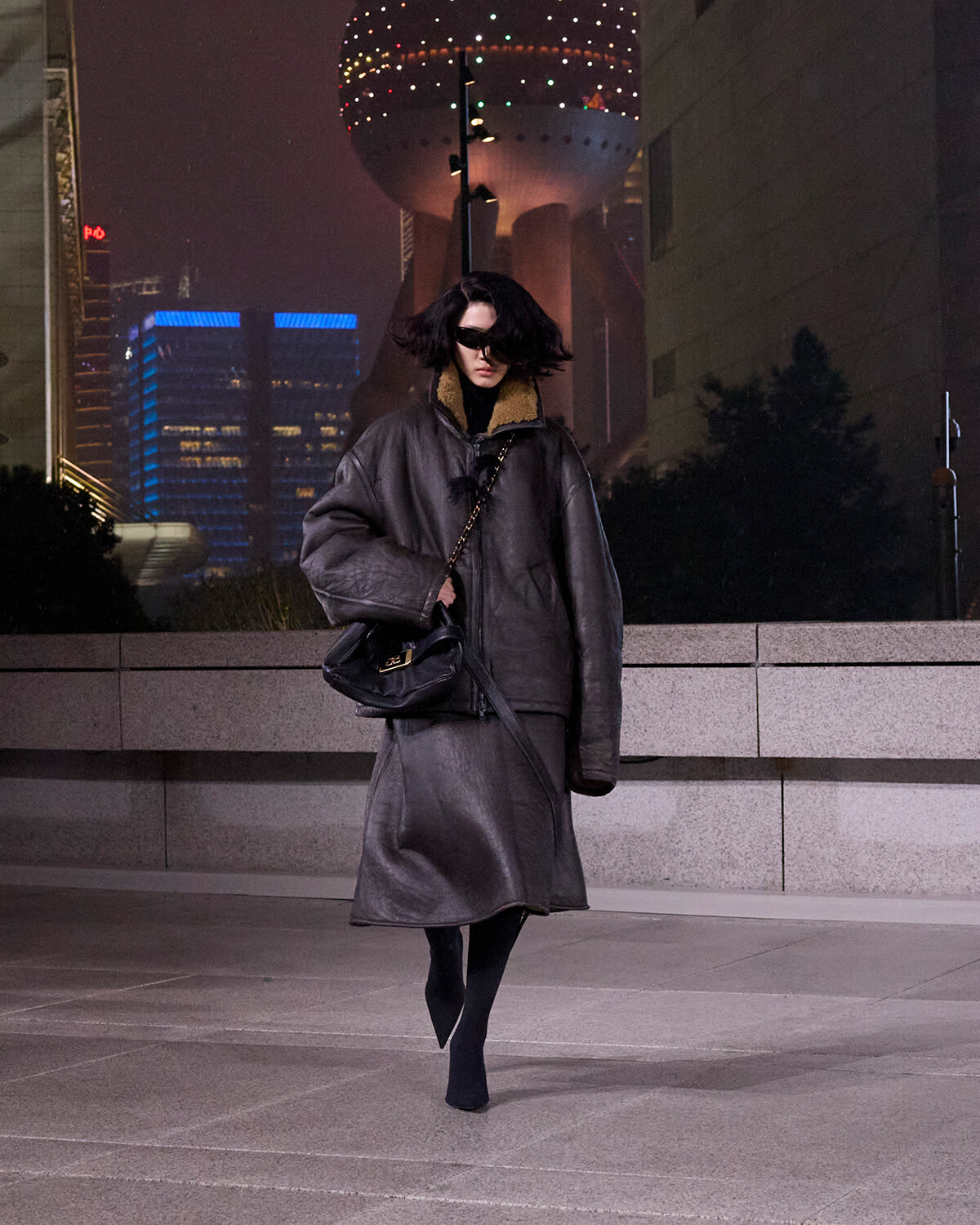 Person in an oversized dark leather coat and sunglasses walks on a city plaza at night, carrying a black shoulder bag.