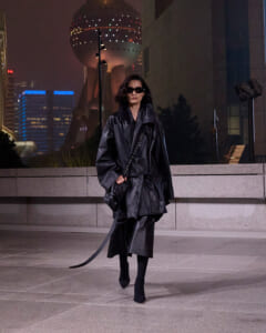 Person in an oversized black leather trench coat and sunglasses walks across a city plaza at night, carrying a small bag against a neon-lit skyline.