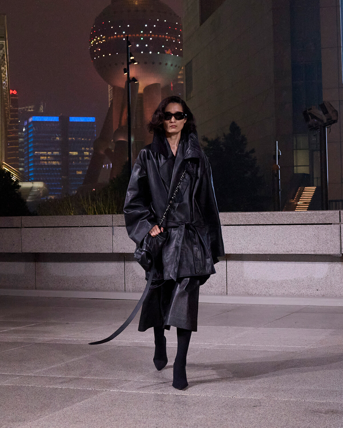 Person in an oversized black leather trench coat and sunglasses walks across a city plaza at night, carrying a small bag against a neon-lit skyline.