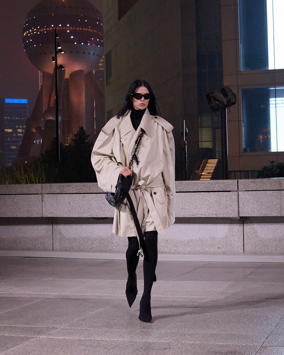 Model walking at night in a beige trench coat, black turtleneck, sunglasses, and knee-high boots, carrying a black bag.