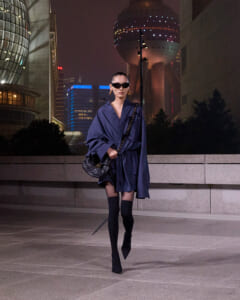 Stylish woman in a navy oversized coat, black thigh-high boots, and sunglasses walking at night in a cityscape.