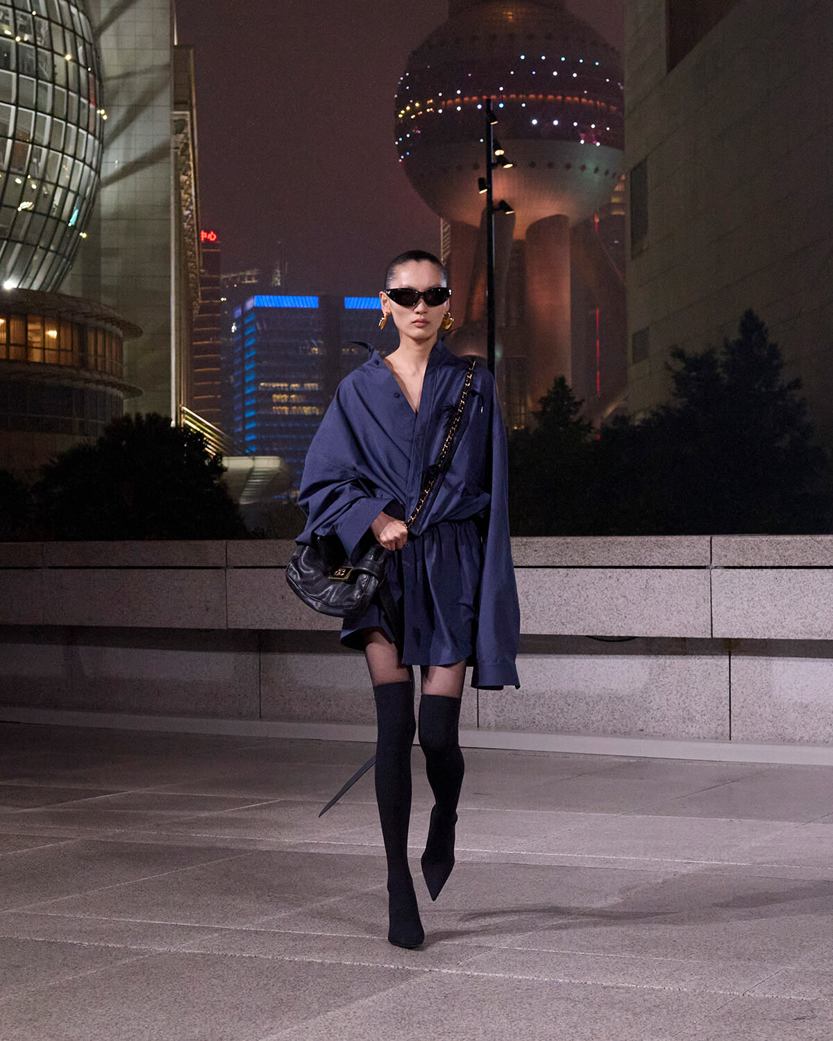 Stylish woman in a navy oversized coat, black thigh-high boots, and sunglasses walking at night in a cityscape.
