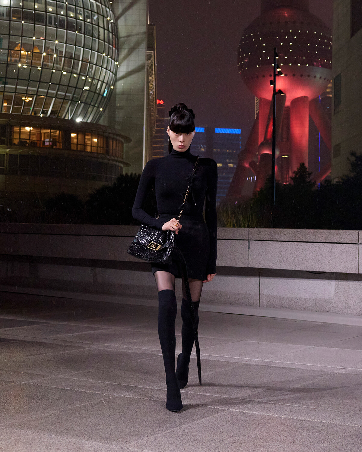 A woman in a black turtleneck dress and thigh-high boots walks at night in a modern city plaza, carrying a quilted black handbag with a chain strap.